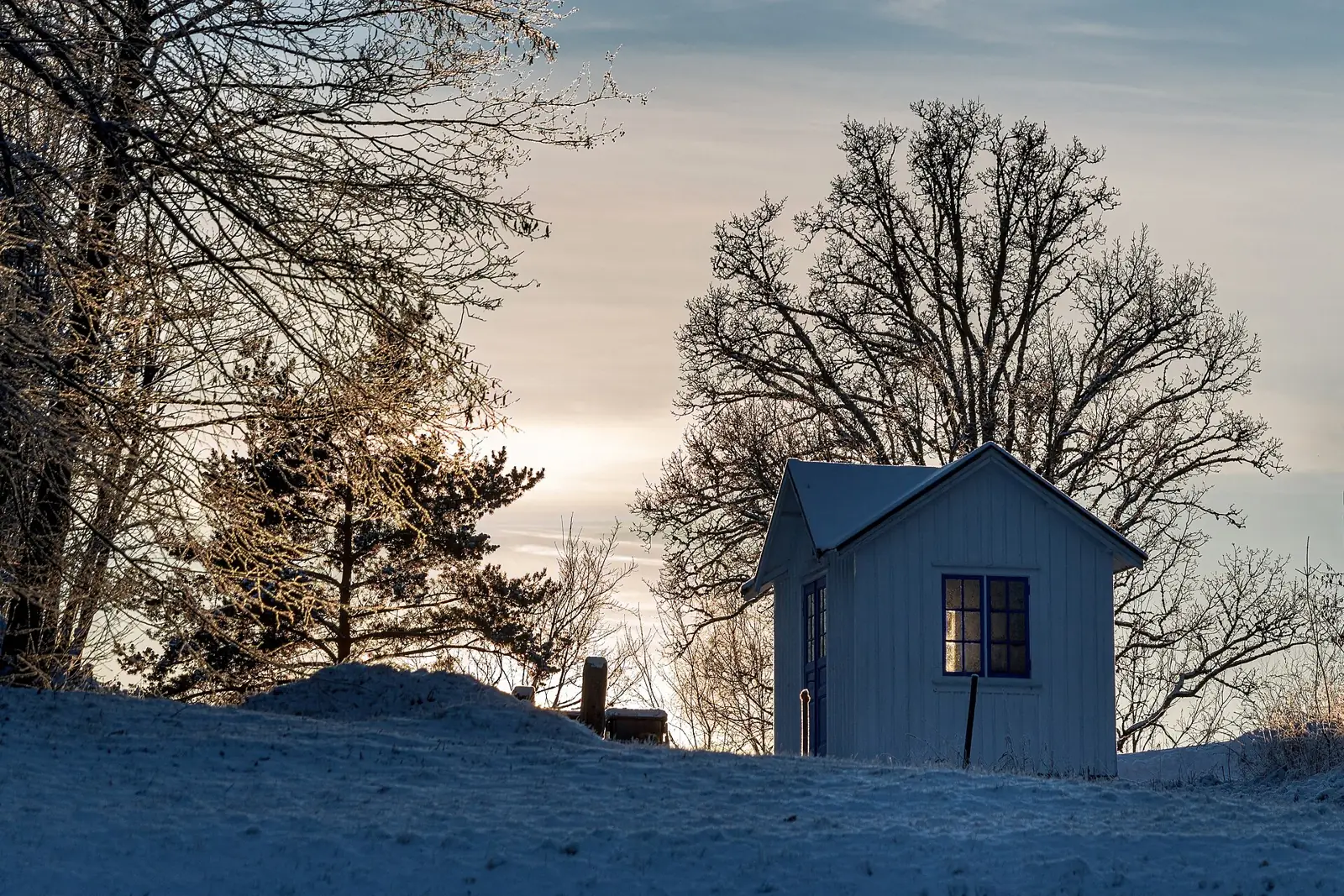 Liten vit stuga på en snöig kulle i lågt vinterljus