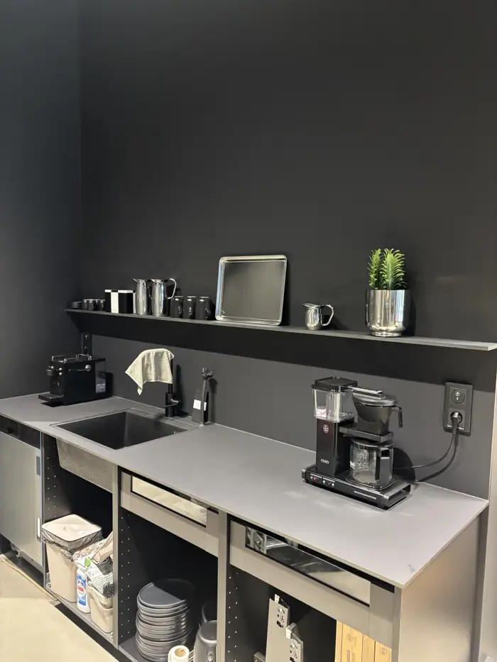 Svarta jordade dubbeluttag ovanför bänkskivan i ett mörkt kontorspentry med kaffebryggare